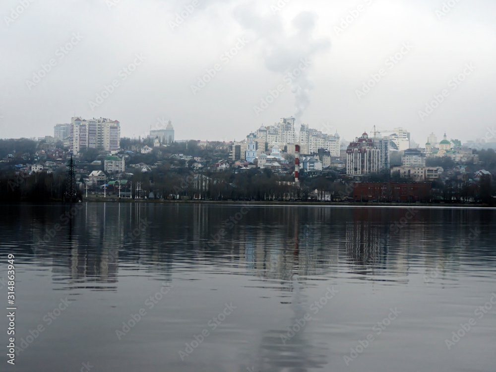 Cloudy winter day in the city. Houses, trees and bridges in the city are reflected in the water of the river on a cloudy day at the beginning of winter.