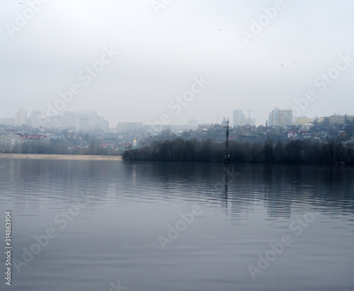 Cloudy winter day in the city. Houses  trees and bridges in the city are reflected in the water of the river on a cloudy day at the beginning of winter.