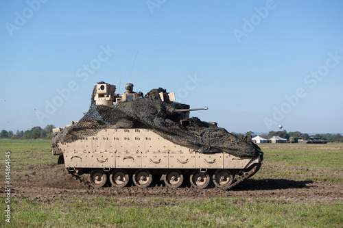 Fighting vehicle used by US army is moving on field and battlefield. Machine has camouflage over turret. Unrecognizable soldier is on the top. Pannign shot with slightly blurred background. photo