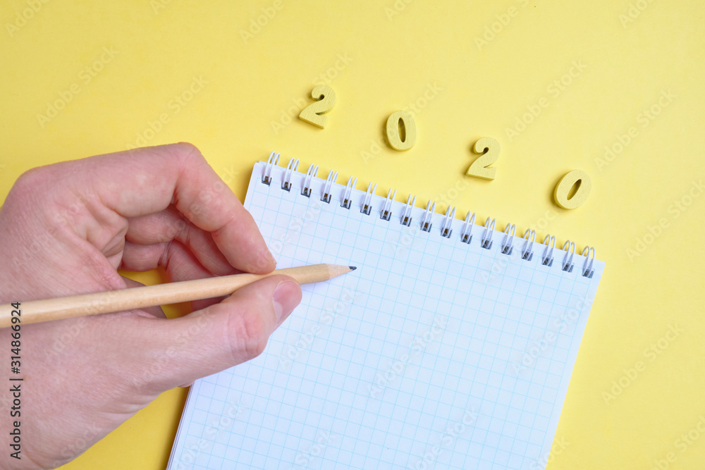 The hand holds a pencil near the notepad next to the wooden letters 2020 on the yellow background. Top view. The hand holds a pencil near the notepad next to the wooden letters 2020 on the yellow background. Top view.