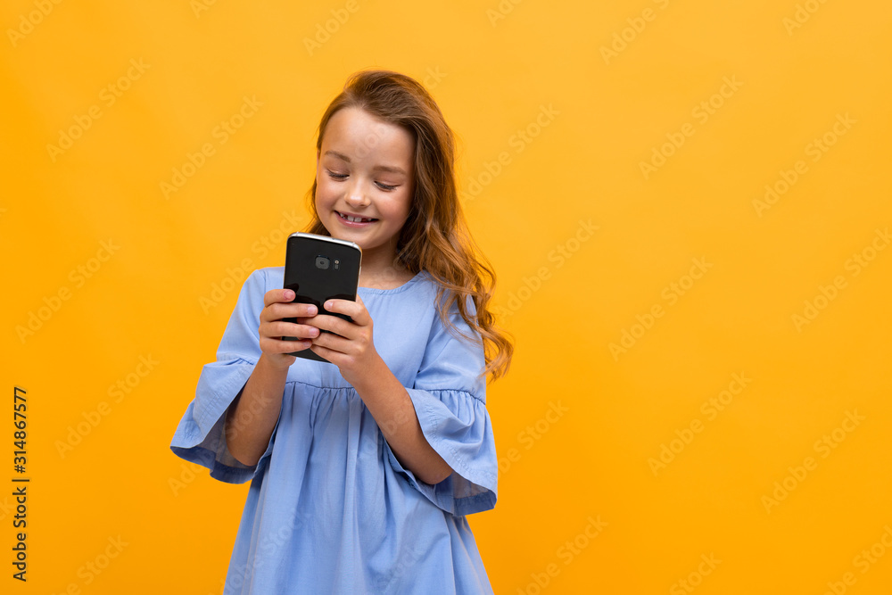 cute smiling girl in a blue dress with a phone in her hands is typing a message on a yellow background with copy space cute smiling girl in a blue dress with a phone in her hands is typing a message on a yellow background with copy space