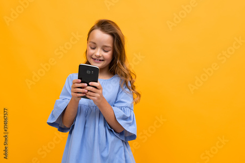 cute smiling girl in a blue dress with a phone in her hands is typing a message on a yellow background with copy space