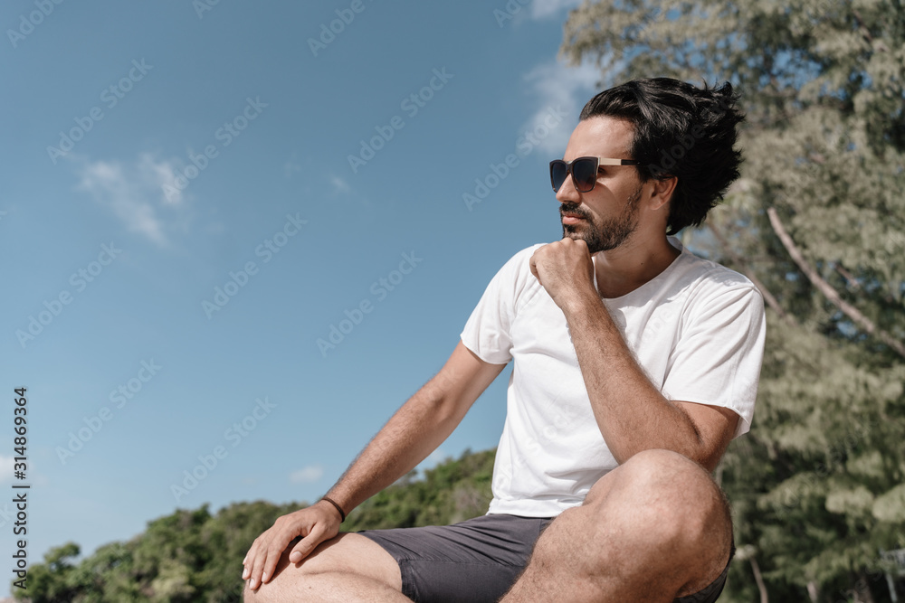 Handsome sexy latin brunette man with a beard in sunglasses and a white T-shirt in profile against a tropical landscape Handsome sexy latin brunette man with a beard in sunglasses and a white T-shirt in profile against a tropical landscape