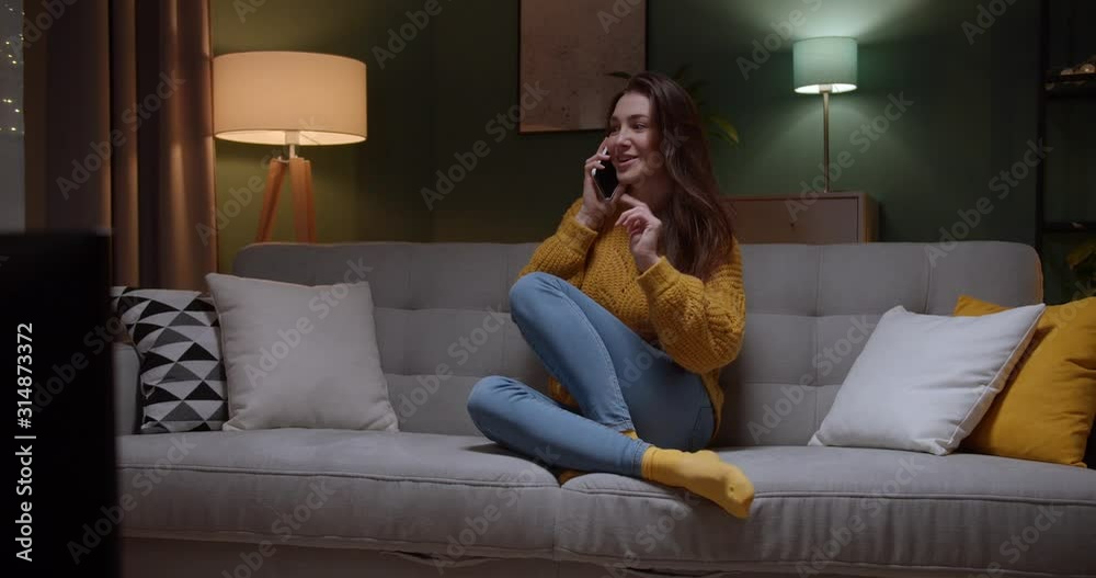 Charming and beautiful young Caucasian girl sitting on the sofa in the living room at night and speaking joyfully on the mobile phone. Inside. Charming and beautiful young Caucasian girl sitting on the sofa in the living room at night and speaking joyfully on the mobile phone. Inside.