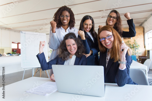 Cheerful businesswomen celebrating success. Happy multiethnic female colleagues raising fists and using laptop in office. Winner gesture concept © Mangostar