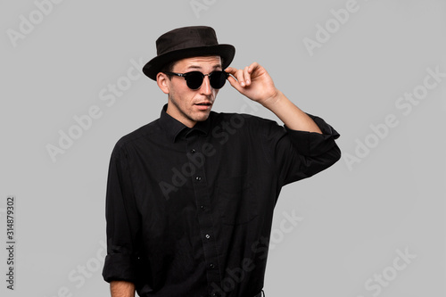 Portrait of a man in a black shirt, pork pie and sun glasses hat isolated over grey background. © ianachyrva