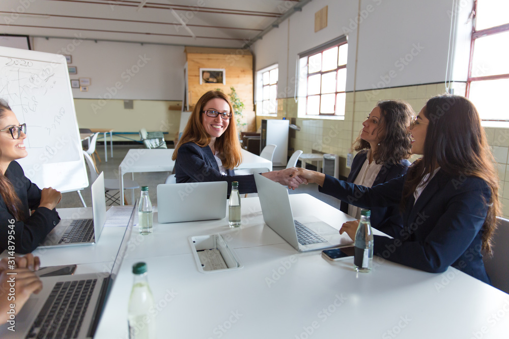 Content businesswomen shaking hands during meeting. Multiethnic female colleagues sitting around table with laptops and discussing work in office. Women in business concept
