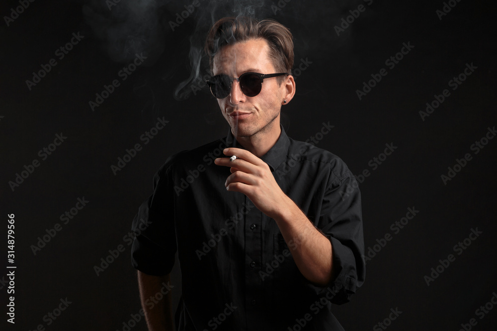 Stylish young man in a black t-shirt and sun glasses smoking a cigarette over black background. Stylish young man in a black t-shirt and sun glasses smoking a cigarette over black background.