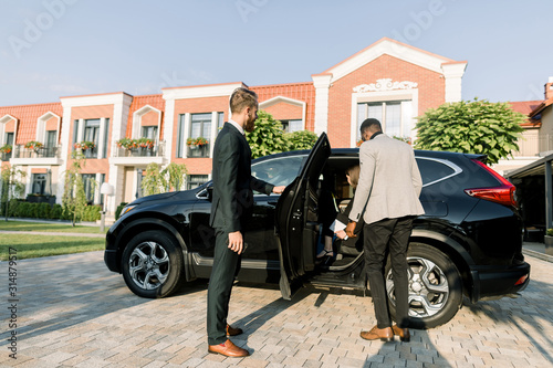 Young Caucasian business man in suit opening black car door for his colleagues, African man and Caucasian woman. Outdoors, business center buildings