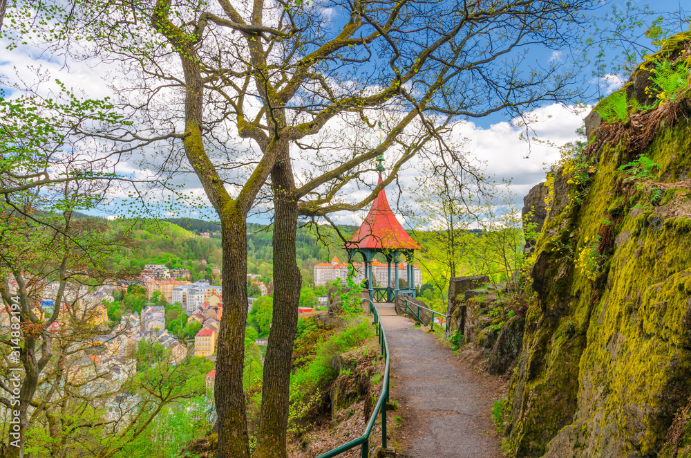 Mayer’s Gloriette wooden gazebo at Deer Jump Jeleni Skok Lookout with top aerial view of Karlovy Vary (Carlsbad) historical city centre, path with trees, big stone rock, West Bohemia, Czech Republic