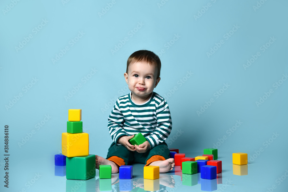 Little baby boy in stylish casual clothing barefoot sitting on floor, playing with colorful toy cubes, showing tongue Little baby boy in stylish casual clothing barefoot sitting on floor, playing with colorful toy cubes, showing tongue