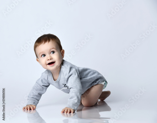 Little baby boy toddler in grey casual jumpsuit and barefoot crawling on floor, smiling and looking up over white wall background