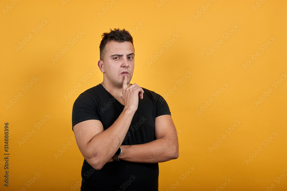 Handsome caucasian young man wearing black shirt isolated on orange background in studio keeping finger on mouth, hugging his stomach. People sincere emotions, lifestyle concept. Handsome caucasian young man wearing black shirt isolated on orange background in studio keeping finger on mouth, hugging his stomach. People sincere emotions, lifestyle concept.