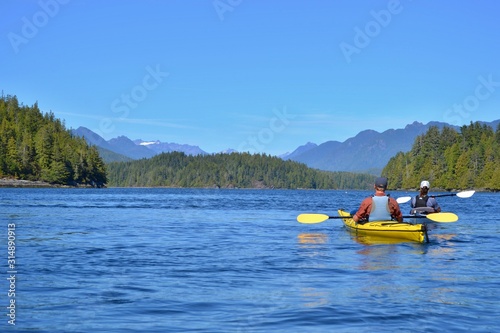 Group of friends on sea kayak in Pacific Ocean near Vancouver Island. Colorful kayaks, trees on the island, man with west, hat and paddle. Blue sky. 