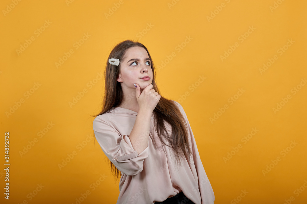 Serious young woman keeping finger on chin, thinking about issue wearing fashion pastel shirt isolated on orange background in studio. People sincere emotions, lifestyle concept. Serious young woman keeping finger on chin, thinking about issue wearing fashion pastel shirt isolated on orange background in studio. People sincere emotions, lifestyle concept.
