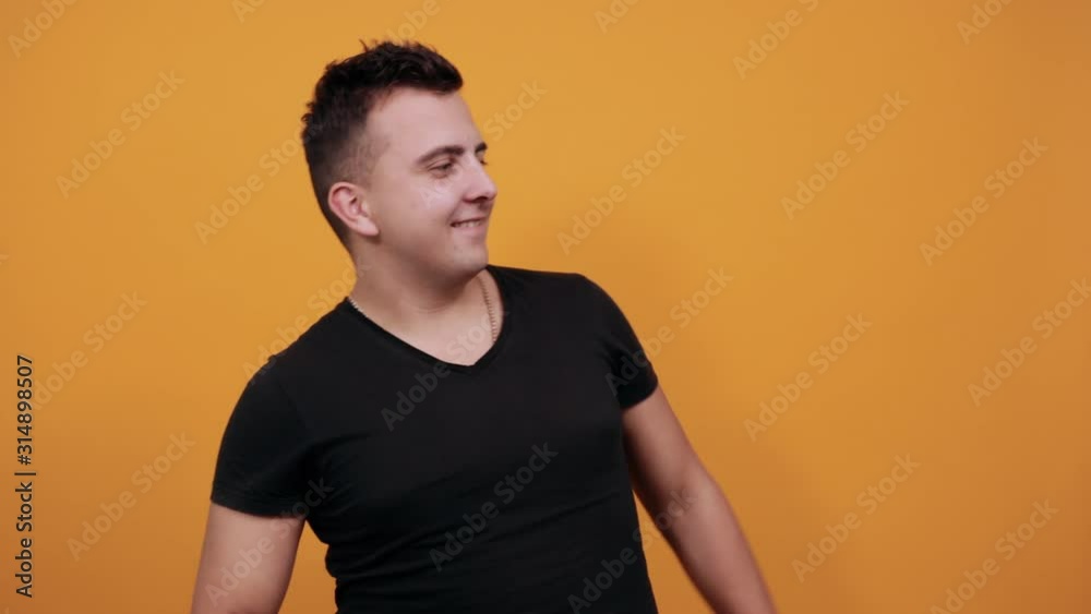 Handsome young man wearing black shirt isolated on orange background in studio smiling, pointing finger aside, keeping hand on neck. People sincere emotions, lifestyle concept. Handsome young man wearing black shirt isolated on orange background in studio smiling, pointing finger aside, keeping hand on neck. People sincere emotions, lifestyle concept.