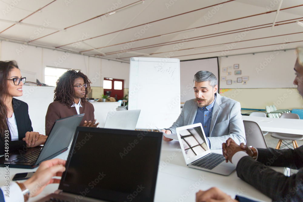Confident leader holding paper document and talking. Office workers sitting at table and listening boss. Business meeting concept Confident leader holding paper document and talking. Office workers sitting at table and listening boss. Business meeting concept