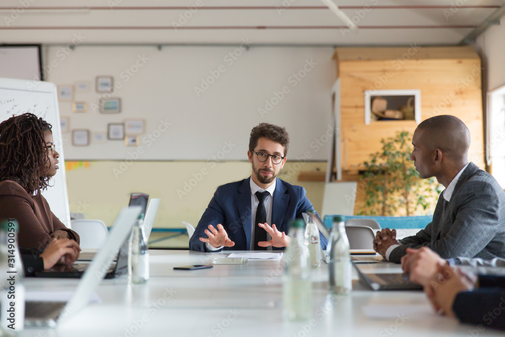 Fototapeta premium Focused project manager communicating with workers. Thoughtful employees sitting at table during morning briefing. Business meeting concept