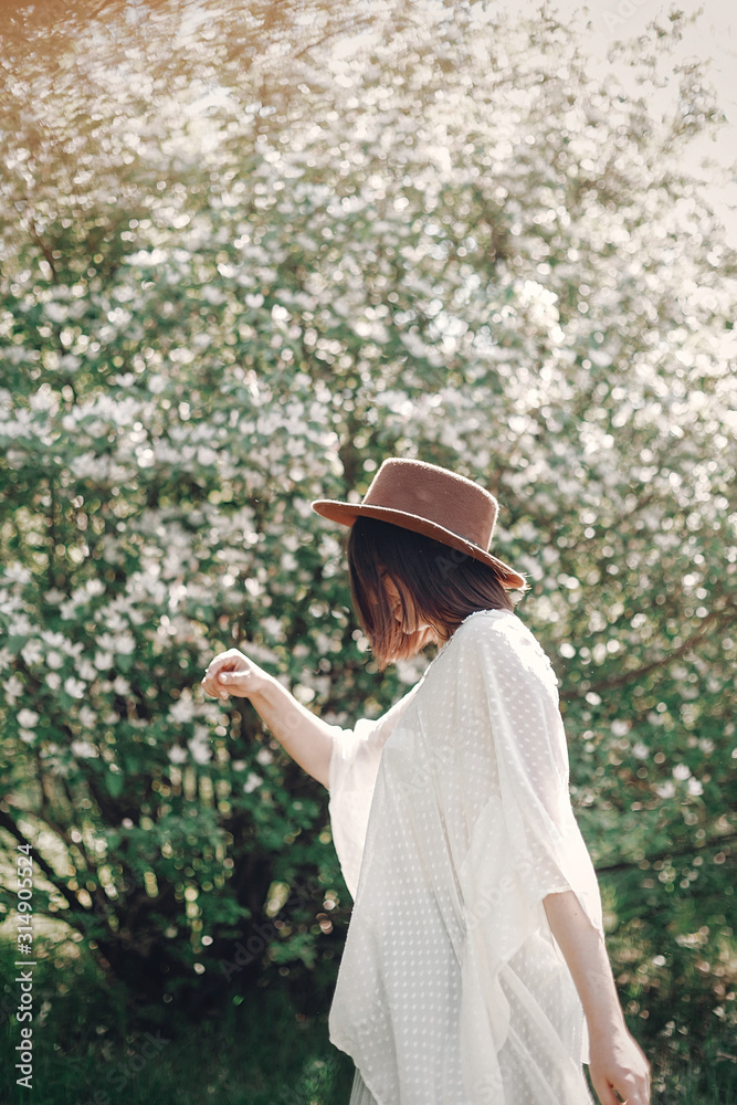 Happy boho woman in hat dancing in sunny light near blooming tree with white flowers in spring park. Stylish hipster girl enjoying spring and moving, having fun. Atmospheric moment