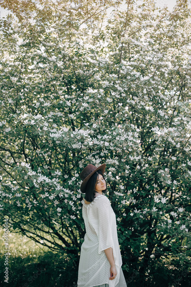 Stylish boho woman in hat posing in blooming tree with white flowers in sunny spring park. Calm portrait of beautiful hipster girl standing in white blooms in spring