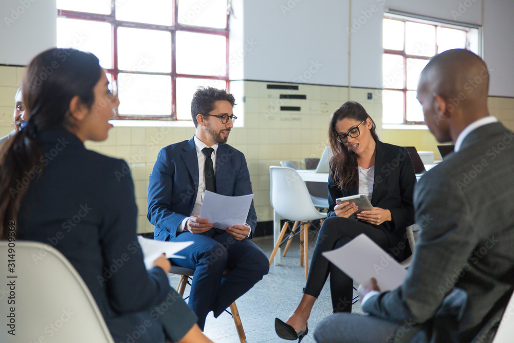 Cheerful managers sitting in chairs with documents and tablet. Confident smiling office employees talking. Teambuilding concept Cheerful managers sitting in chairs with documents and tablet. Confident smiling office employees talking. Teambuilding concept