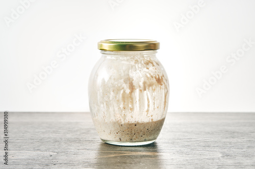 Homemade sourdough starter for bread in a glass jar with closed lid on a table against white background. Made of whole wheat flour. Bubbling fermentation, messy streaks inside container