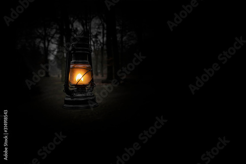 Vintage kerosene oil lantern lamp burning with a soft glow light in an dark forest / wood. Light in the darkness. Blurred background image of a glowing lantern against dark night time. Old lamp.