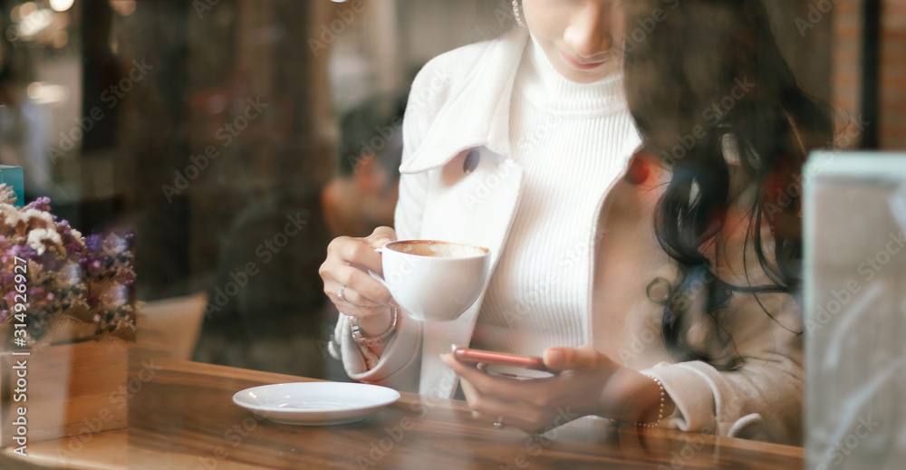 Shot photo through window glass. Businesswoman relaxing and holding coffee to drink while she using mobile phone in cafe Shot photo through window glass. Businesswoman relaxing and holding coffee to drink while she using mobile phone in cafe