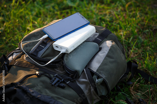 Using a power Bank to charge your phone and gadgets in the field. White portable battery charges on a backpack, on a stump, on a tourist Mat.