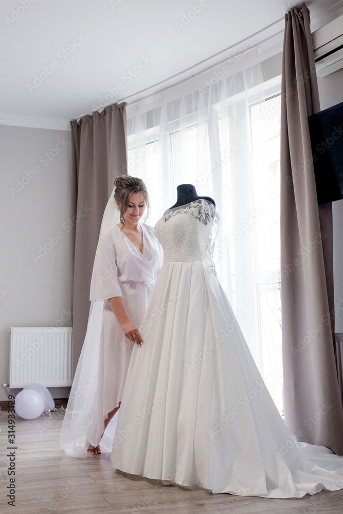 Young woman in silk bathrobe touching her wedding dress and smiling while standing near the window. Wedding day. Beauty, fashion. Happiest bridal day. Enjoy every moment. Young woman in silk bathrobe touching her wedding dress and smiling while standing near the window. Wedding day. Beauty, fashion. Happiest bridal day. Enjoy every moment.