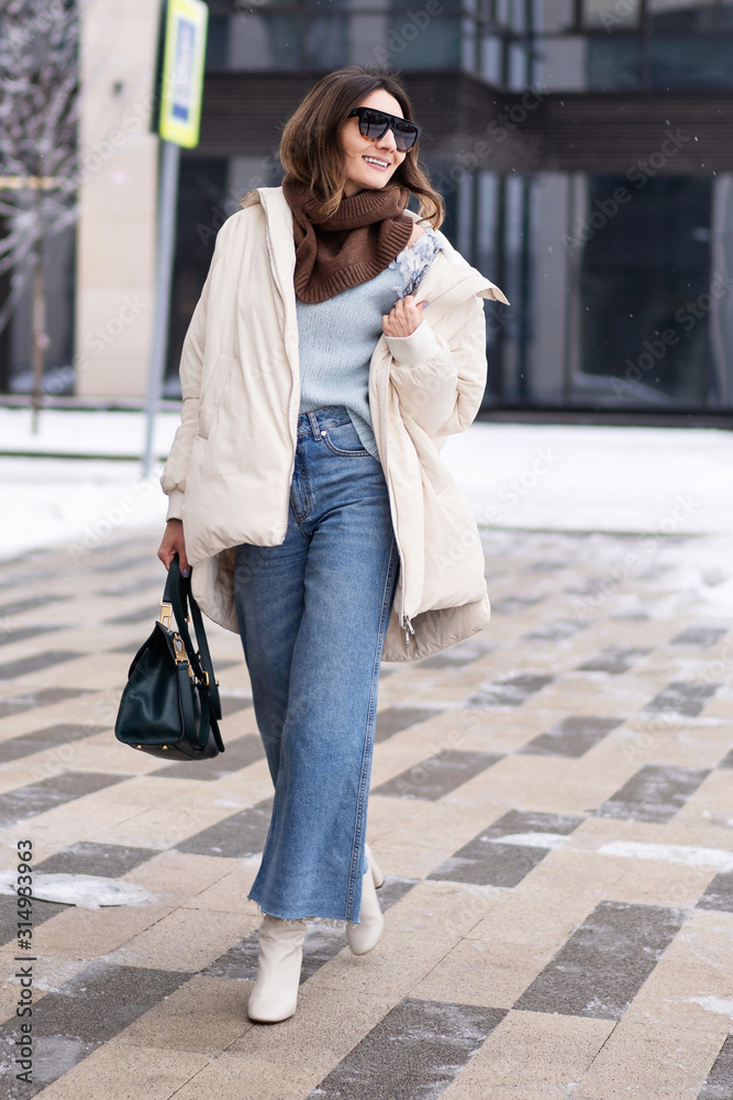 Caucasian model girl in a beige oversized down jacket, knitted sweater, flared jeans with a handbag and glasses, walks in the winter near the office building. Life style Caucasian model girl in a beige oversized down jacket, knitted sweater, flared jeans with a handbag and glasses, walks in the winter near the office building. Life style
