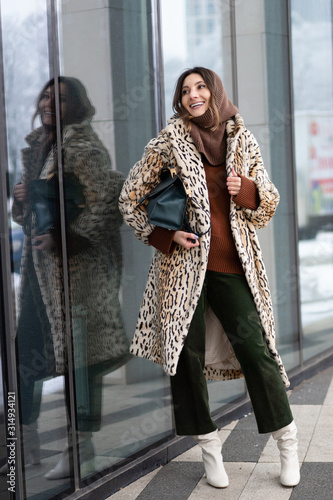 European girl in a leopard fur coat, corduroy pants with a handbag stands near the office building. Life style