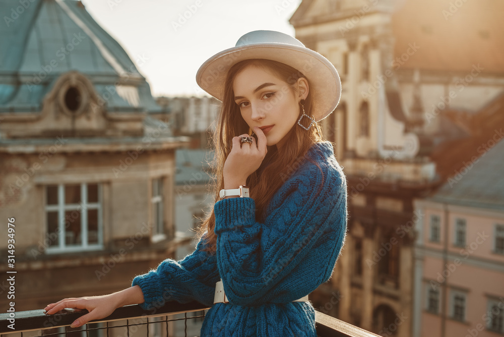 Young elegant fashionable brunette woman, model wearing trendy blue knitted sweater, stylish white hat, wrist watch, earrings, ring, posing at sunset, in European city. Copy, empty space for text - obrazy, fototapety, plakaty 