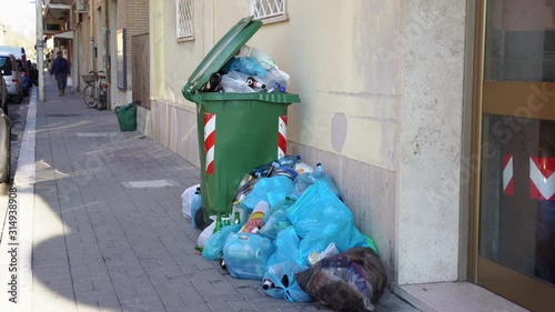 Green plastic container is overfilled with trash and organic waste on the street of Ladispoli, Italy. People throwing out garbage on the ground under the windows of residential building. Trash problem photo