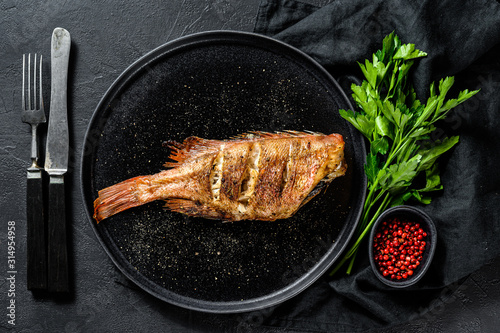 Sea perch baked served on a round plate with parsley. Black background. Top view.