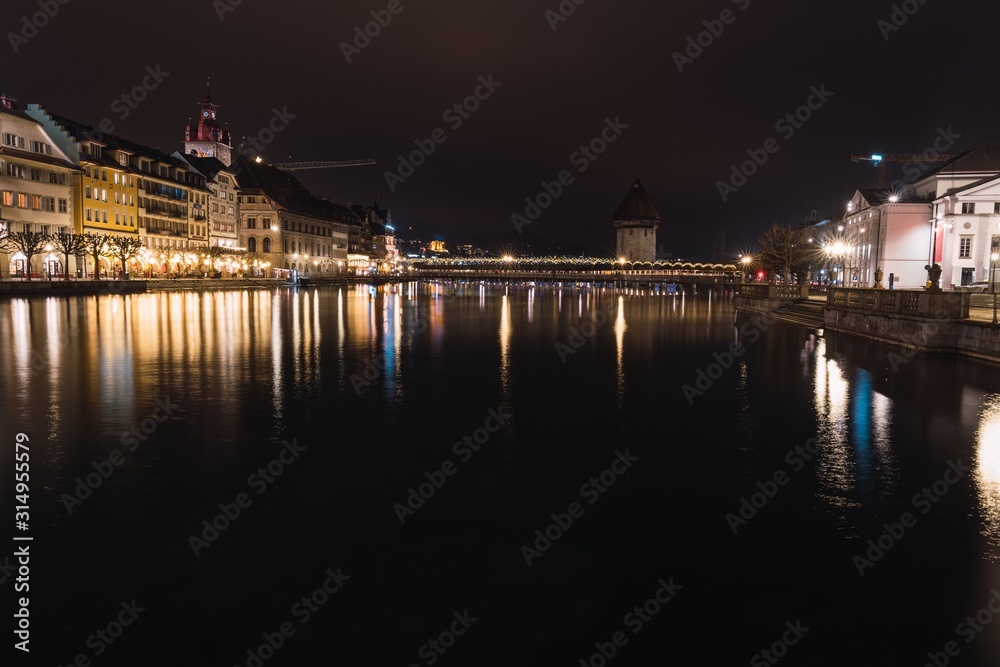 Illuminated buildings surrounding a river with a bridge in the middle of the city at night with the lights reflected in the water