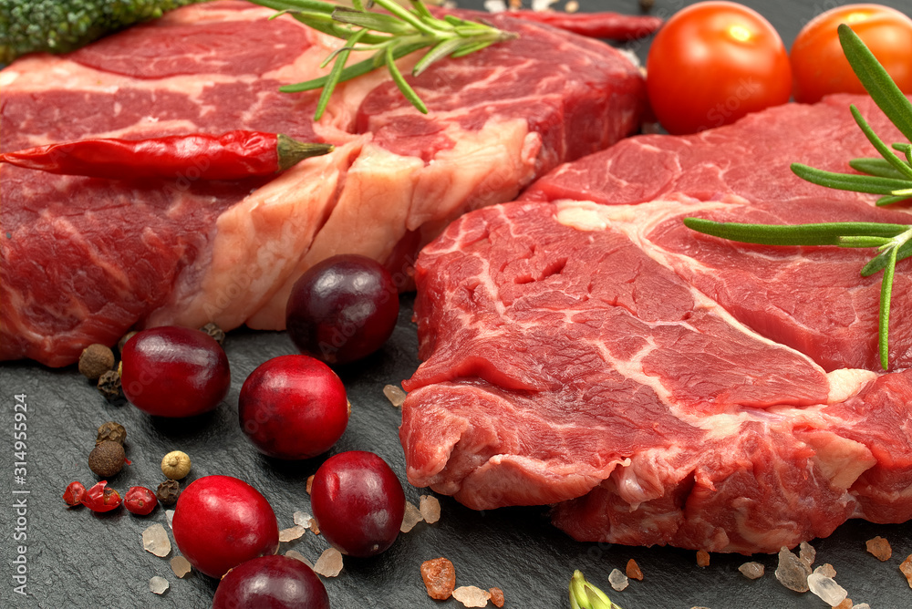 Raw rib eye steak with spices and vegetables. Ingredients for restaurant meal. Fresh meat, salt, rosemary, thyme, chilli, cherry tomatoes, garlic on black stone. Food background.