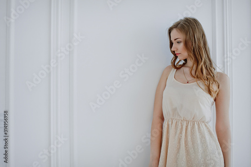 beautiful young girl in beige dress with long blond hair stands near white wall. girl waiting for gift by March 8. young woman without flowers on international women's day.
