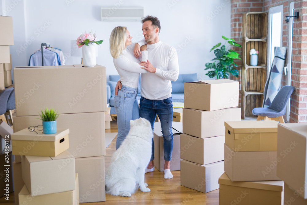 Fototapeta premium Young beautiful couple with dog standing at new home around cardboard boxes
