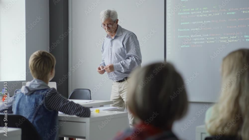 Pan left view of old teacher with pen standing by projector screen and explaining computer science and coding basics to kids in school class Pan left view of old teacher with pen standing by projector screen and explaining computer science and coding basics to kids in school class