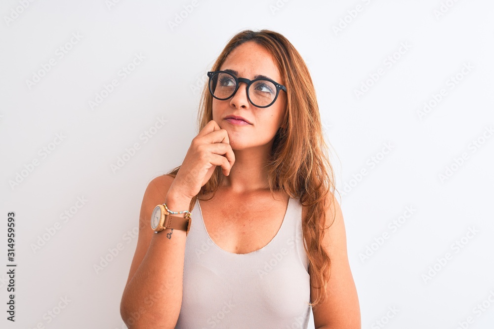 Young redhead woman wearing glasses standing over white isolated background with hand on chin thinking about question, pensive expression. Smiling with thoughtful face. Doubt concept. Young redhead woman wearing glasses standing over white isolated background with hand on chin thinking about question, pensive expression. Smiling with thoughtful face. Doubt concept.