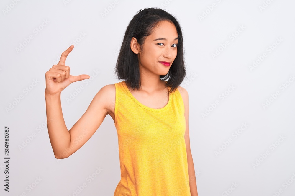 Young chinese woman wearing yellow casual t-shirt standing over isolated white background smiling and confident gesturing with hand doing small size sign with fingers looking and the camera. Measure Young chinese woman wearing yellow casual t-shirt standing over isolated white background smiling and confident gesturing with hand doing small size sign with fingers looking and the camera. Measure