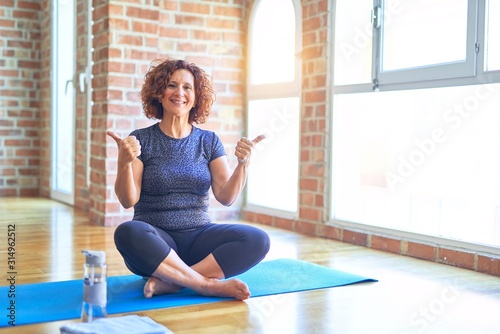 Middle age beautiful sportswoman wearing sportswear sitting on mat practicing yoga at home success sign doing positive gesture with hand, thumbs up smiling and happy. Cheerful expression and winner