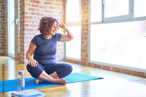 Middle age beautiful sportswoman wearing sportswear sitting on mat practicing yoga at home very happy and smiling looking far away with hand over head. Searching concept.
