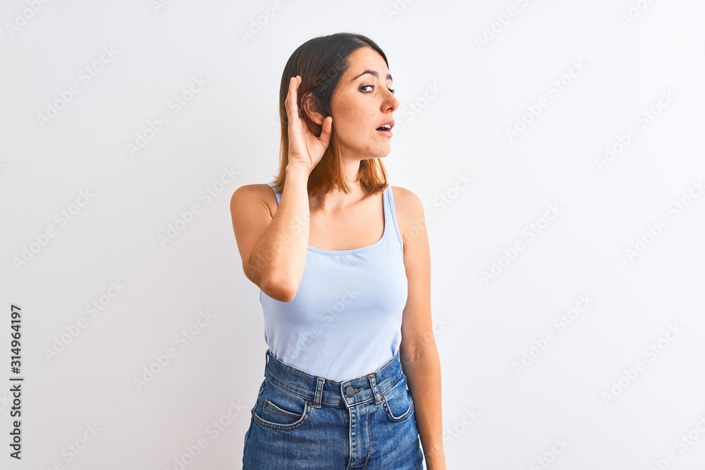 Beautiful redhead woman standing over isolated background smiling with hand over ear listening an hearing to rumor or gossip. Deafness concept.