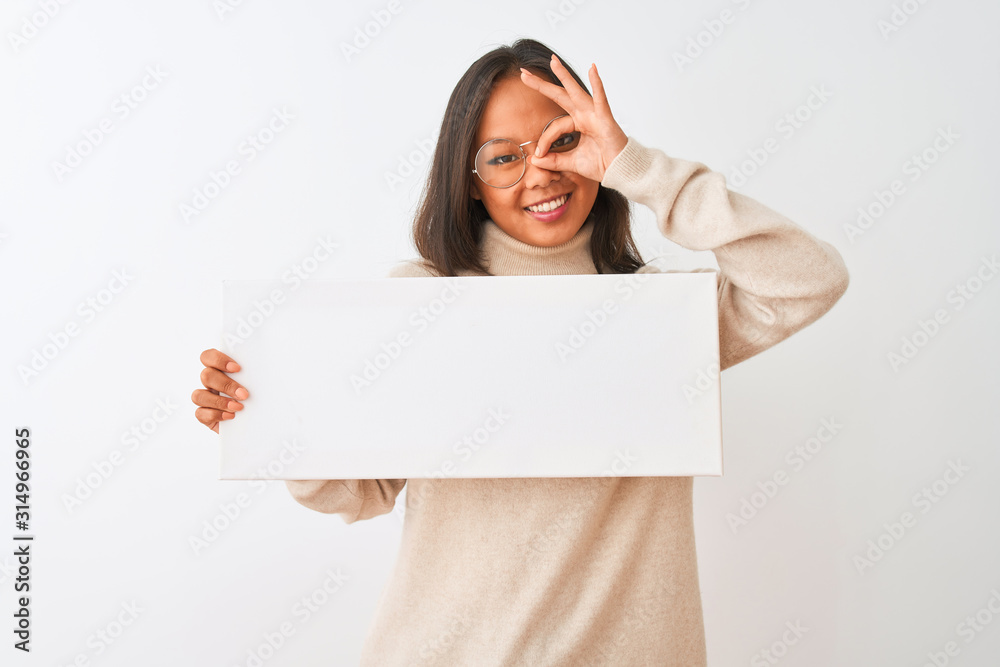 Young beautiful chinese woman wearing glasses holding banner over isolated white background with happy face smiling doing ok sign with hand on eye looking through fingers Young beautiful chinese woman wearing glasses holding banner over isolated white background with happy face smiling doing ok sign with hand on eye looking through fingers