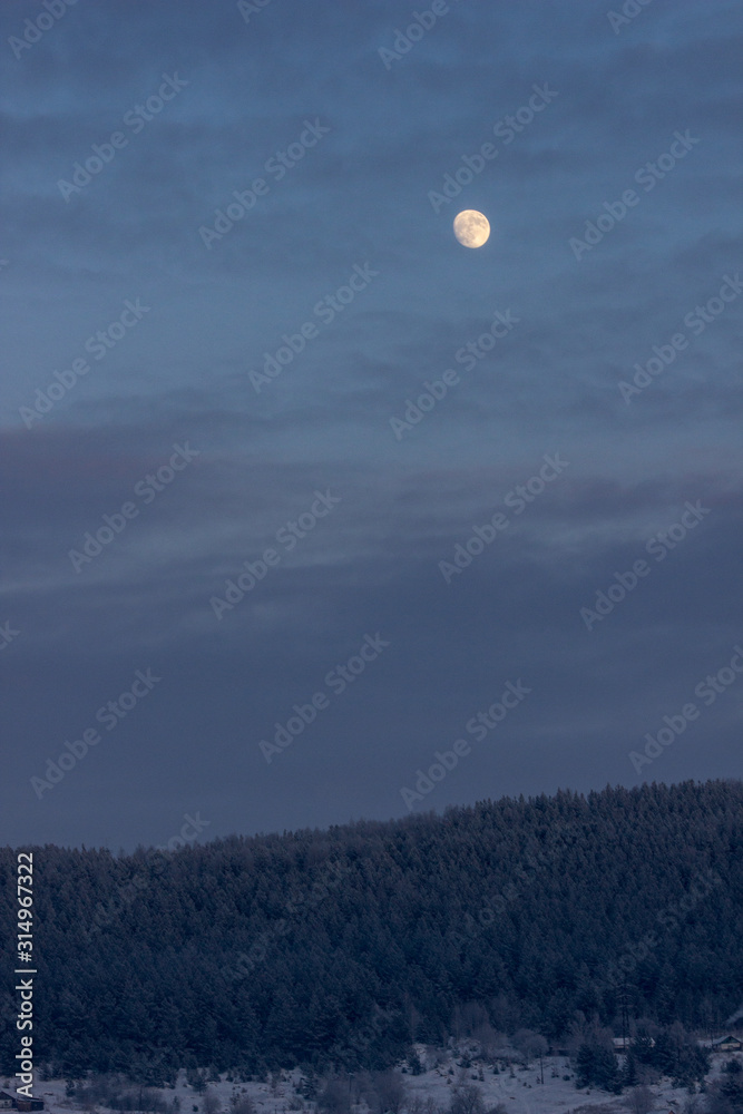 Full moon in the sky with clouds over the evening, winter forest. Dark sky with clouds, a round moon and coniferous forest on top of a snowy mountain. Forest silhouette in hoarfrost. Full moon in the sky with clouds over the evening, winter forest. Dark sky with clouds, a round moon and coniferous forest on top of a snowy mountain. Forest silhouette in hoarfrost.