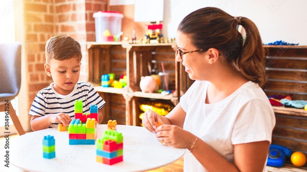 Fototapeta premium Beautiful teacher and toddler boy playing with construction blocks bulding tower at kindergarten