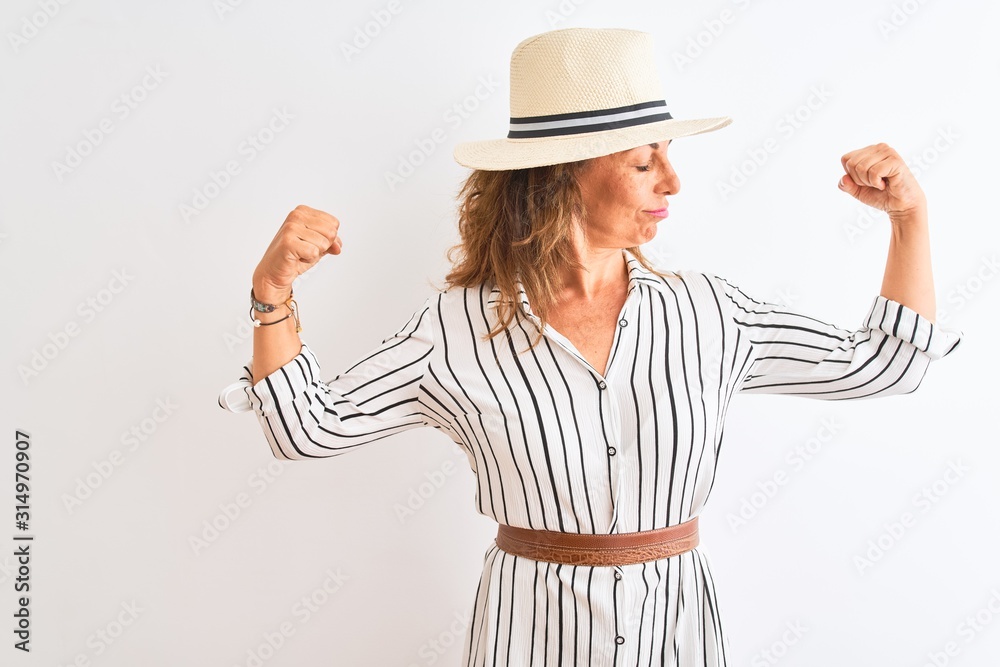 Middle age businesswoman wearing striped dress and hat over isolated white background showing arms muscles smiling proud. Fitness concept. Middle age businesswoman wearing striped dress and hat over isolated white background showing arms muscles smiling proud. Fitness concept.