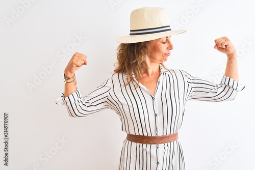 Middle age businesswoman wearing striped dress and hat over isolated white background showing arms muscles smiling proud. Fitness concept.